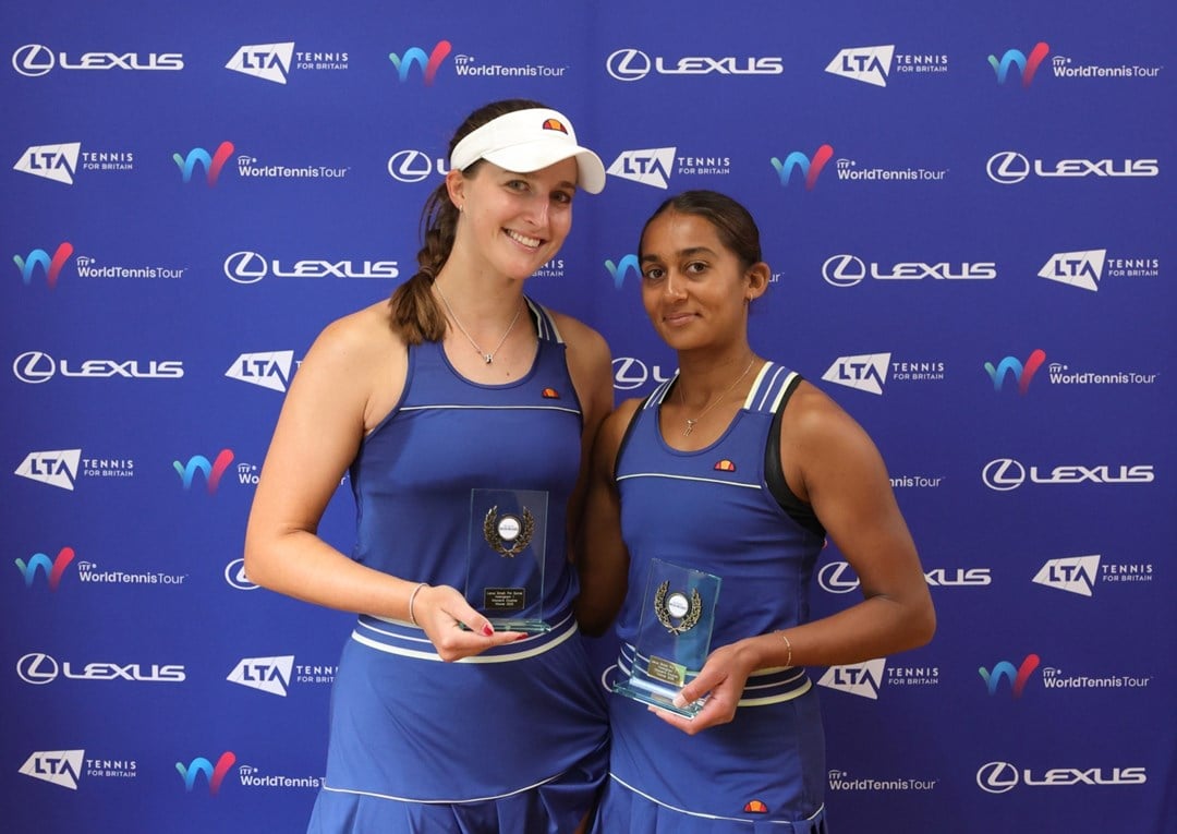 Two women wearing blue tennis dresses and holding glass trophies, stood in front of a blue background with sponsor logos from the LTA, ITF World Tennis Tour, and Lexus on display