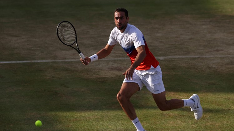 A man runs to hit a tennis ball on a grass tennis court. He is holding a tennis racket in his right hand, and is wearing white shoes, white shorts, and a t-shirt with a white top half and red bottom half
