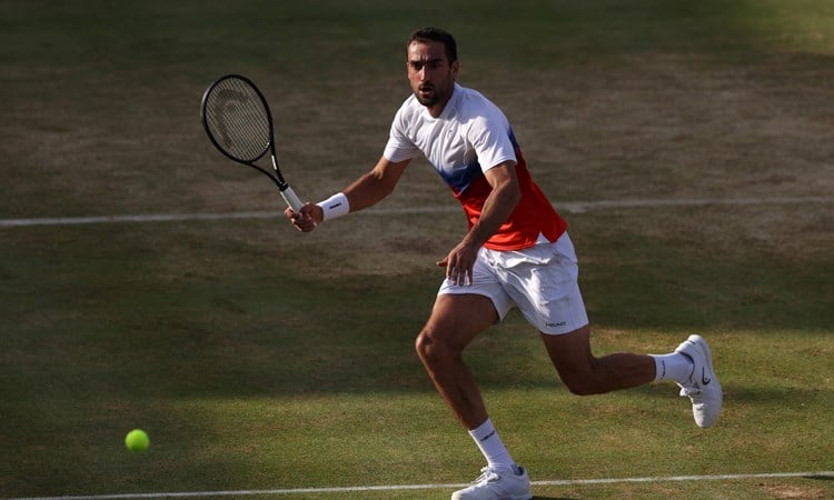 A man runs to hit a tennis ball on a grass tennis court. He is holding a tennis racket in his right hand, and is wearing white shoes, white shorts, and a t-shirt with a white top half and red bottom half