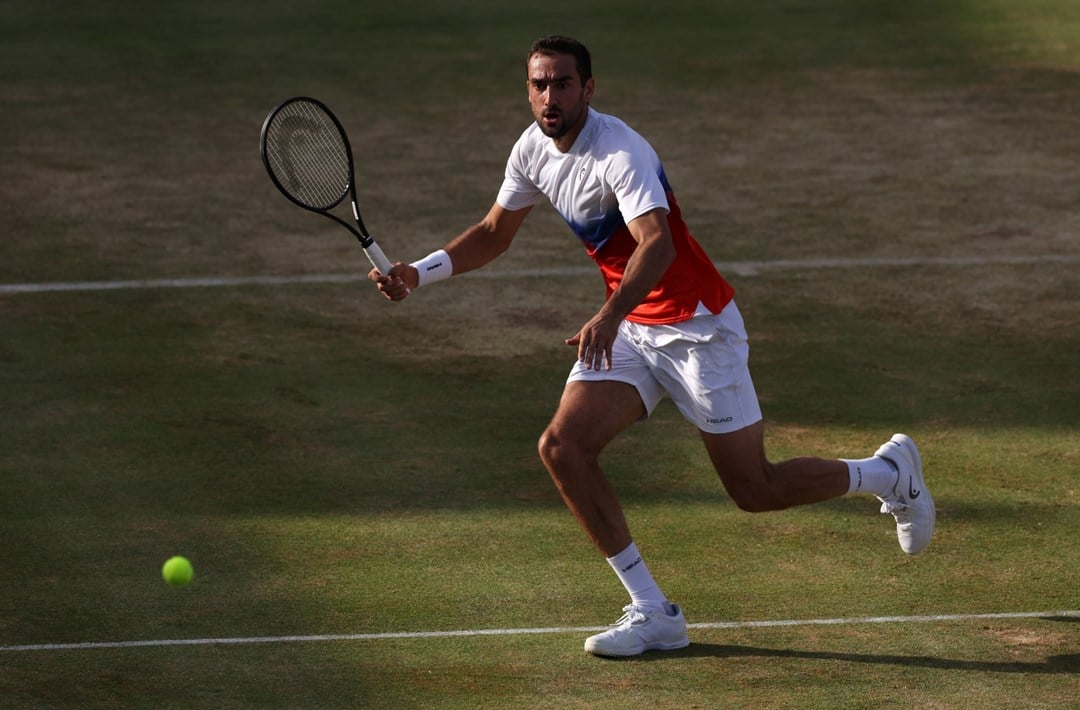 A man runs to hit a tennis ball on a grass tennis court. He is holding a tennis racket in his right hand, and is wearing white shoes, white shorts, and a t-shirt with a white top half and red bottom half