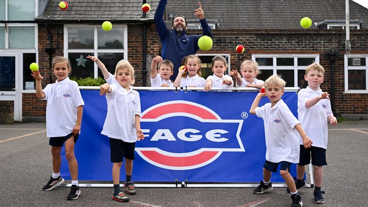 A group of children, stood in front and behind of a  blue banner with the word 'FAGE' on it, throw tennis balls towards the camera. There is an adult man stood with the group as well.
