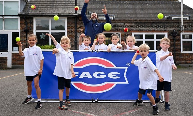 A group of children, stood in front and behind of a  blue banner with the word 'FAGE' on it, throw tennis balls towards the camera. There is an adult man stood with the group as well.