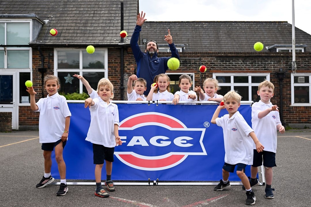 A group of children, stood in front and behind of a blue banner with the word 'FAGE' on it, throw tennis balls towards the camera. There is an adult man stood with the group as well.