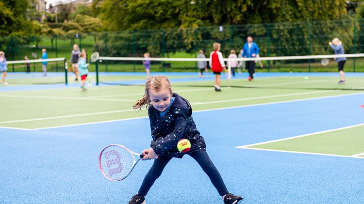 A young girl preparing to hit a ball with her tennis racket surrounded by other children on a park tennis court