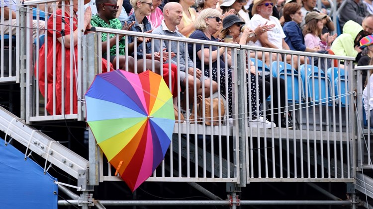 A multi-coloured umbrella is open, in the shape of a heart, resting in front of railing, with spectators sat behind