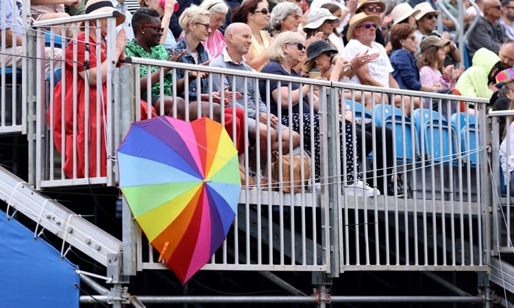 A multi-coloured umbrella is open, in the shape of a heart, resting in front of railing, with spectators sat behind