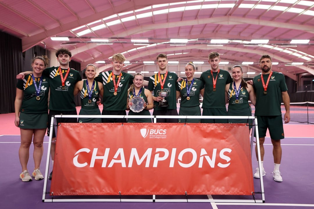 A group of men and women wearing green sports kit and with medals around their necks stand in front of a large red banner with the word 'Champions' written on it