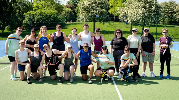 A group of woman stood together on a tennis court, smiling at the camera, with blue sky and greenery in the background