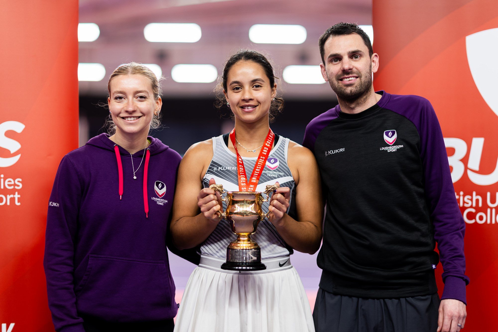 A girl in grey sports kit holds a gold trophy and wears a medal round her neck. Two people in purple sports kit, a woman on the left and a ma on the right, stand either side of her
