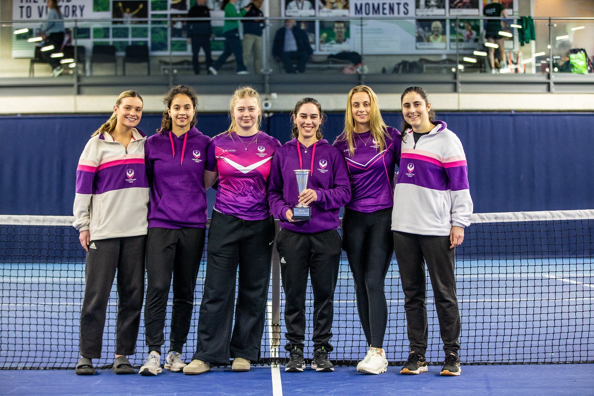A group of women in purple sports kit stand in front of a tennis net, with the woman in the centre left of the group holding a silver trophy