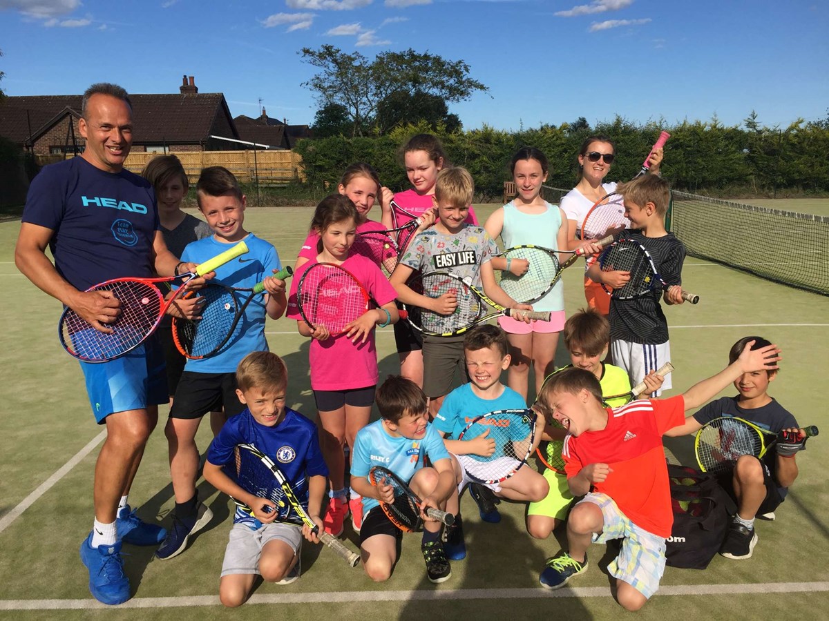 A tennis coach and a group of kids posing and smiling with their tennis rackets on court