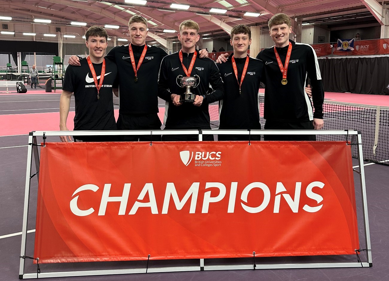 Five men in a line with their arms over each other's shoulders, all wearing black with medals around their necks, stood behind a large red banner that says the word 'champions' on it. The man in the very centre is stood holding a trophy