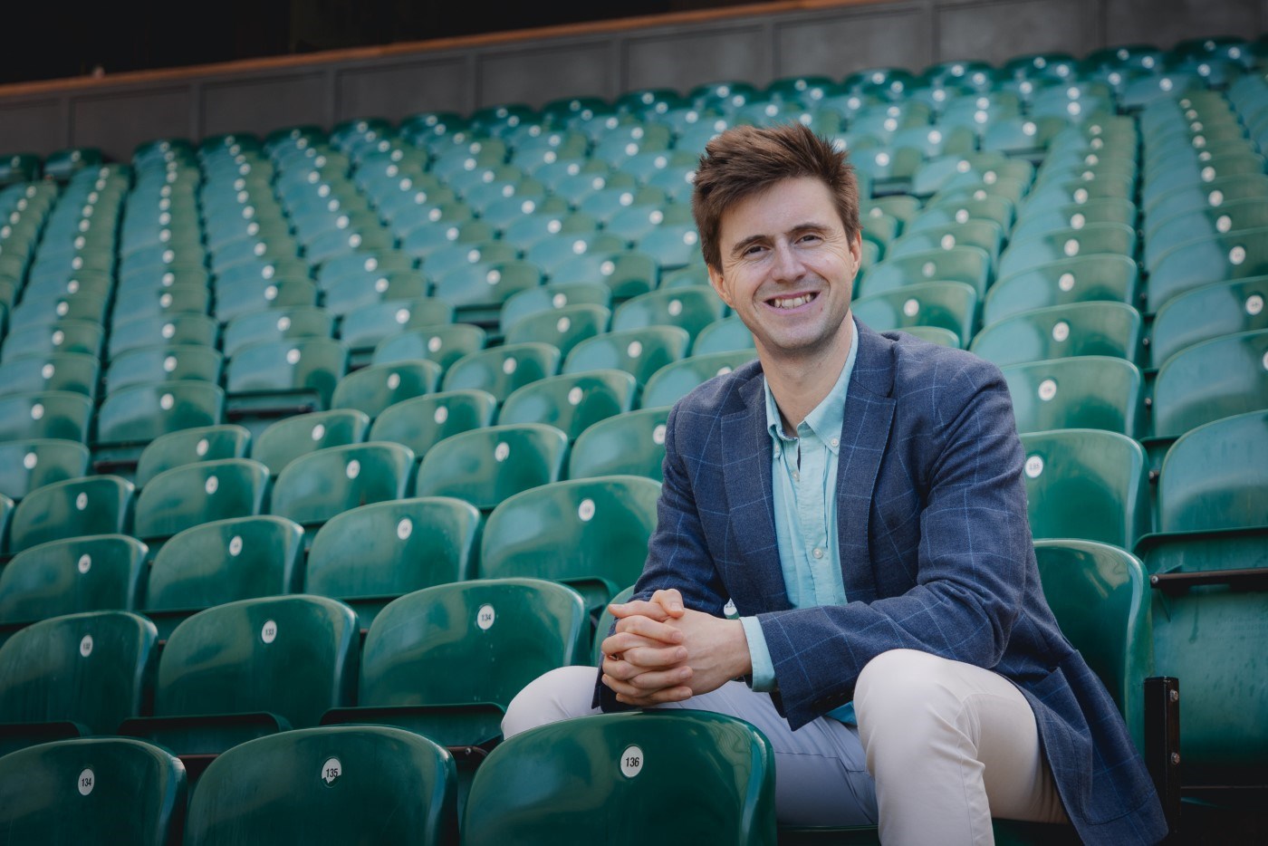 A man sat in green seating, clasping his hands together, wearing a blue checked blazer jacket
