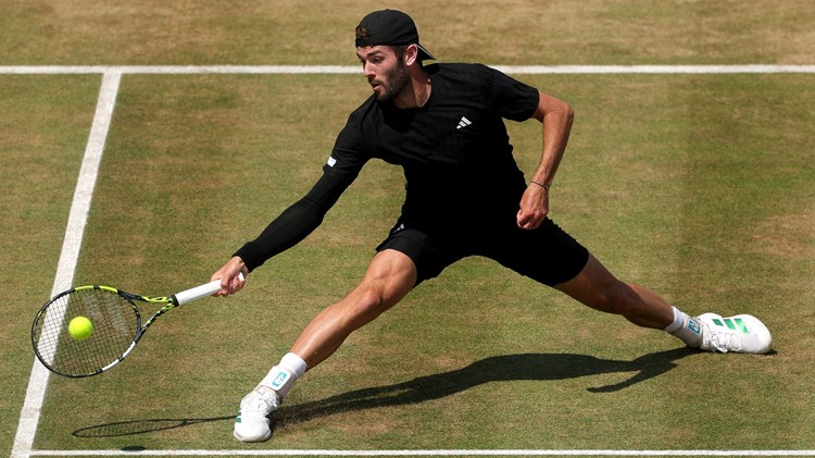 Jacob Fearnley with his legs spread wide, reaching for a ball on a grass court