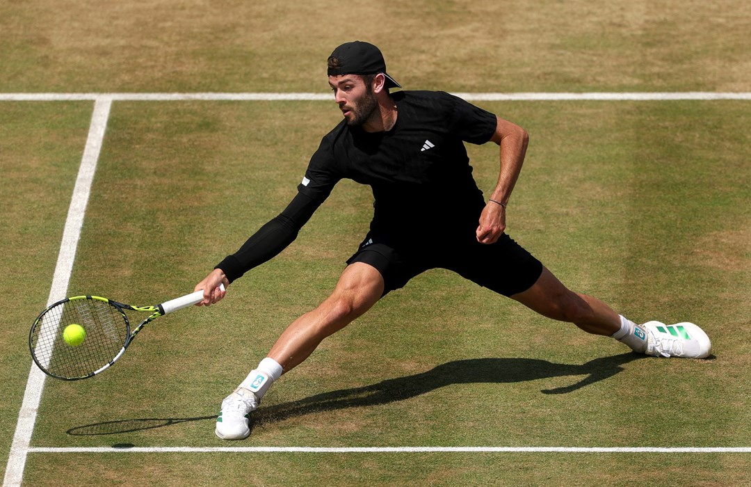 Jacob Fearnley with his legs spread wide, reaching for a ball on a grass court