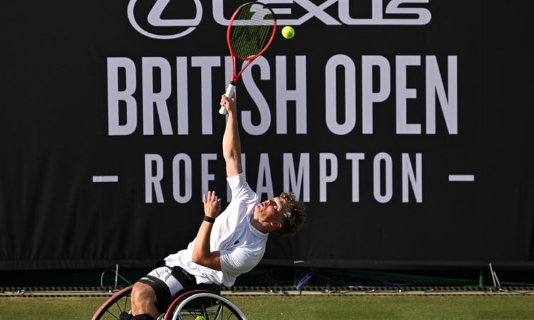 Ben Bartram serves on the grass in front of a Lexus British Open Roehampton sign