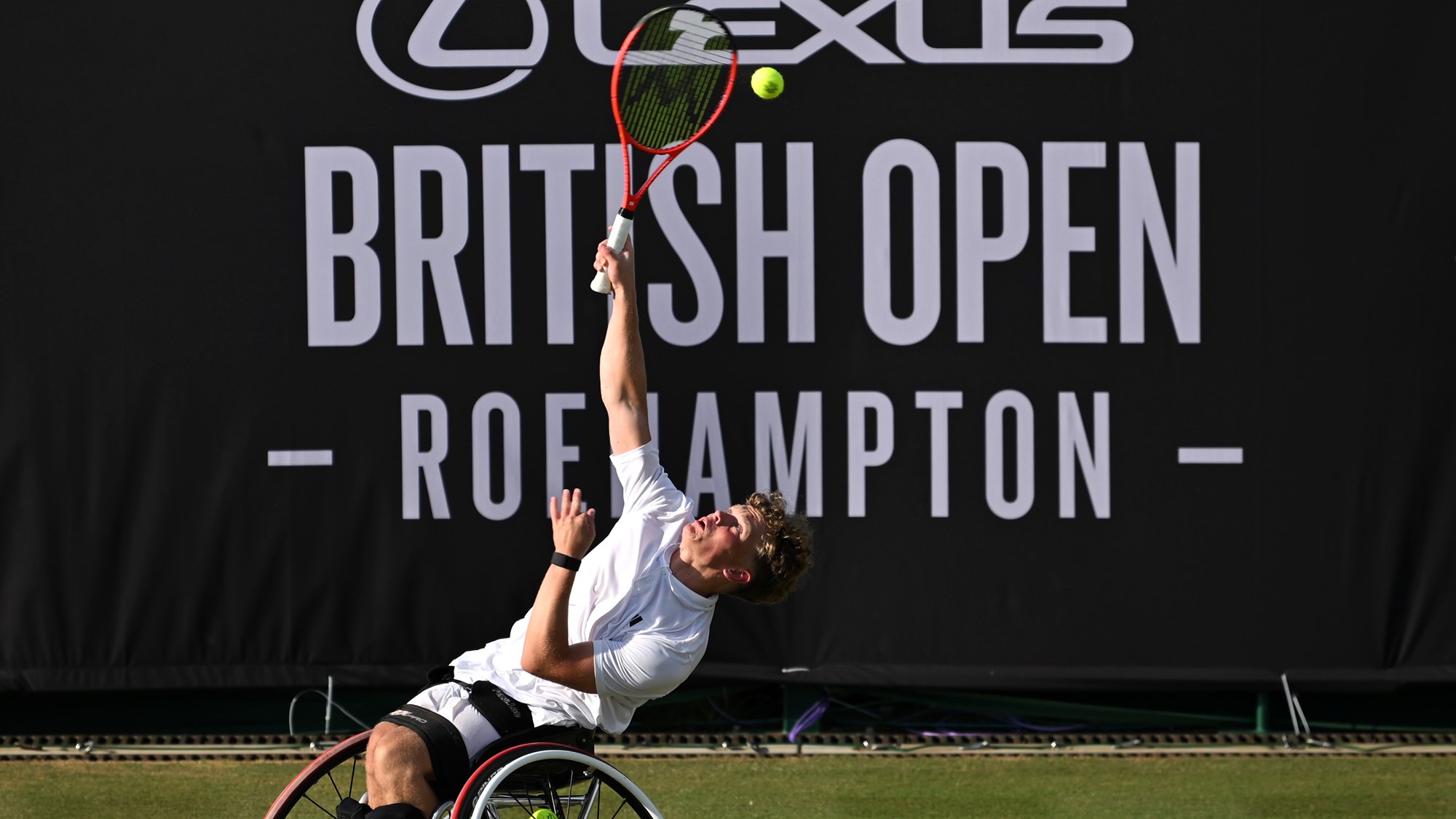 Ben Bartram serves on the grass in front of a Lexus British Open Roehampton sign