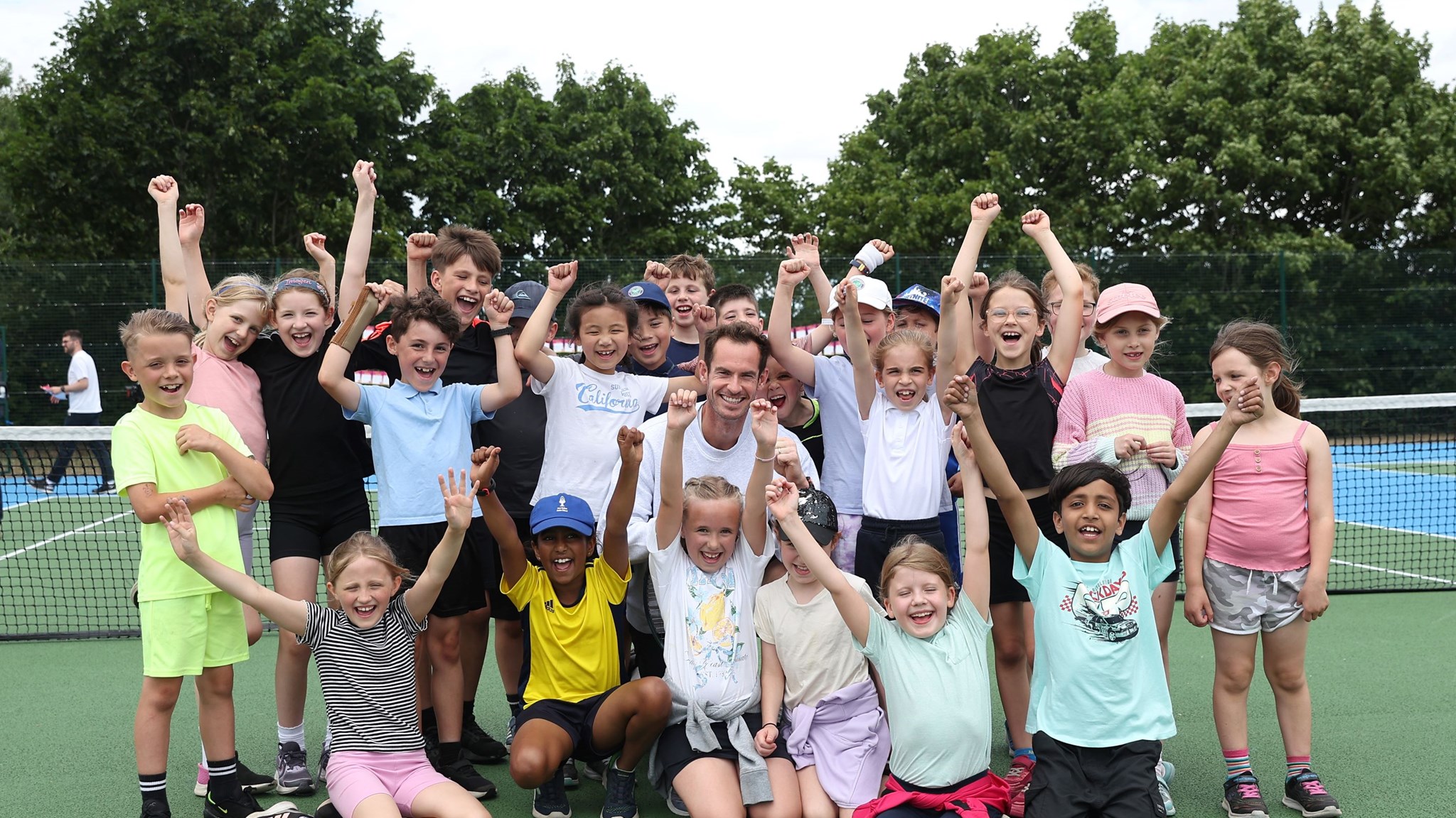 Andy Murray smiling in the middle of a group of young kids cheering with their hands in the air on a park court