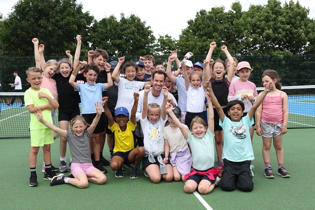 Andy Murray smiling in the middle of a group of young kids cheering with their hands in the air on a park court