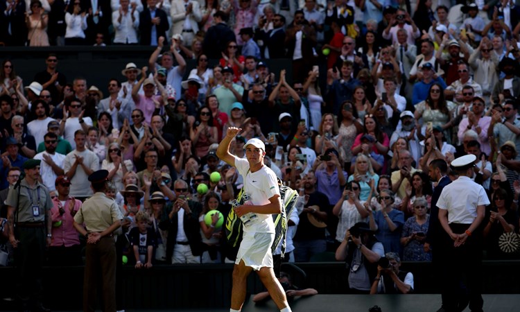 Oliver Tarvet, dressed in white, holding a thumb up to the crowd after his second round match against Carlos Alcaraz at Wimbledon 