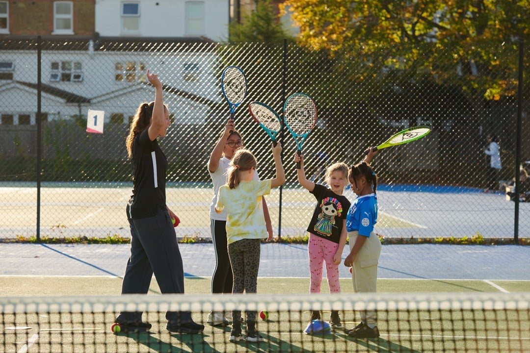 Female coach cheering with hands in the air next to a bunch of kids cheering with their tennis rackets in the air