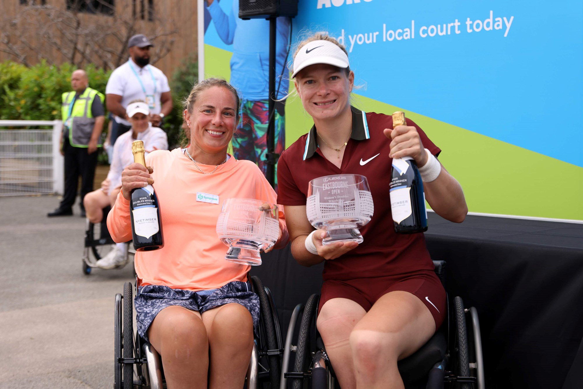 Lucy Shuker and Diede De Groot smiling and holding their trophies and a bottle of Nyetimber champagne after winning the women's wheelchair doubles title at the Lexus Eastbourne Open