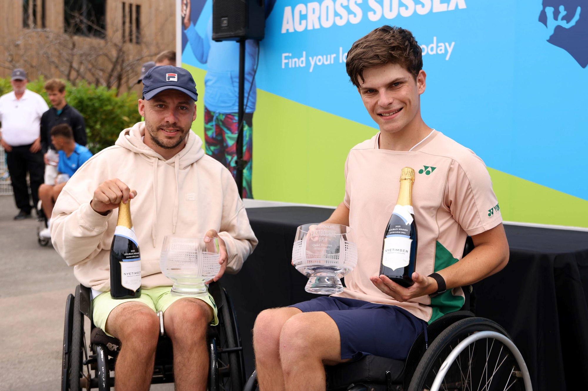 Andy Lapthorne and Greg Slade smiling while holding their quad doubles trophies and a bottle of Nyetimber at the Lexus Eastbourne Open