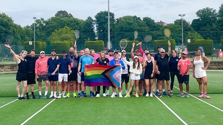 A group of people stand on a tennis court, holding tennis rackets in the air . In the middle of the group three people between them hold a Pride flag