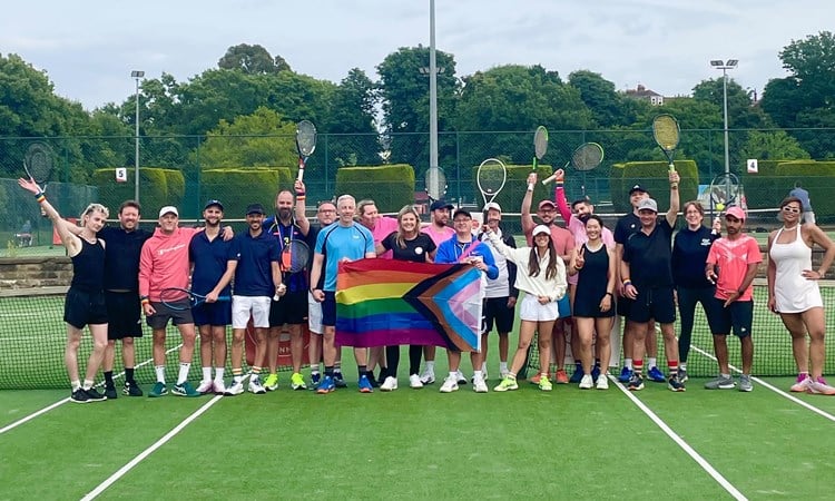 A group of people stand on a tennis court, holding tennis rackets in the air . In the middle of the group three people between them hold a Pride flag