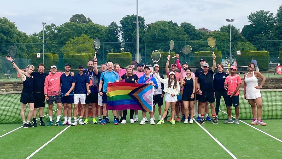 A group of people stand on a tennis court, holding tennis rackets in the air . In the middle of the group three people between them hold a Pride flag