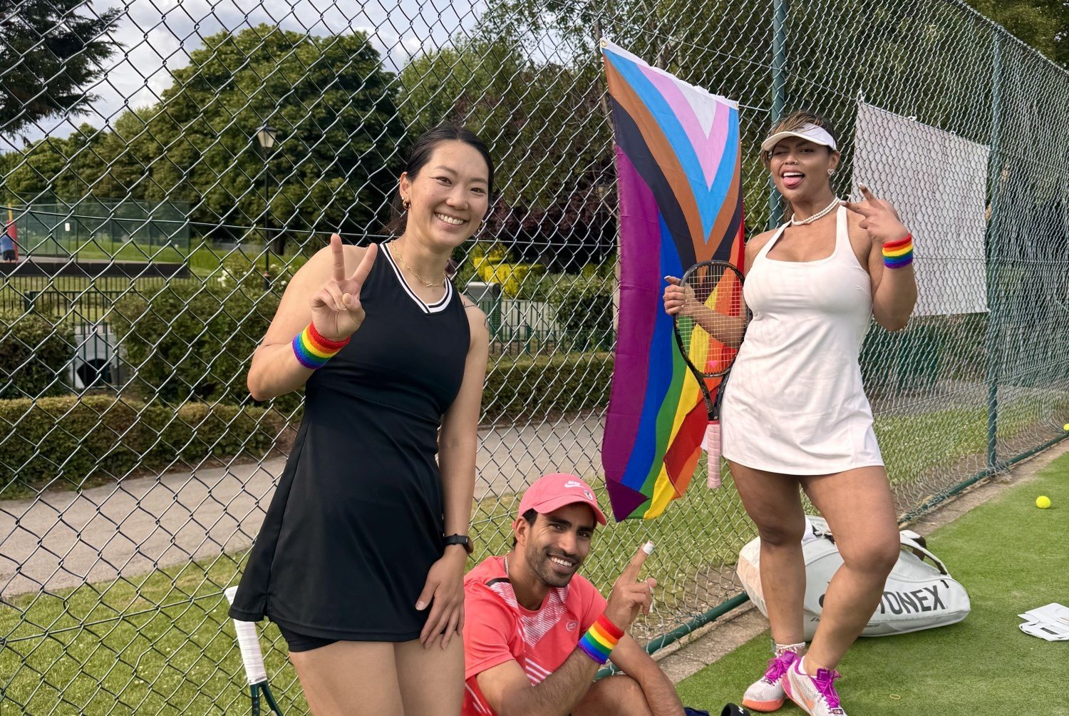 Three people standing smiling at the camera, each wearing rainbow sweatbands on their wrists. The person on the right is also holding a tennis racket, and behind ther group, a brightly coloured flag is attached to the fence
