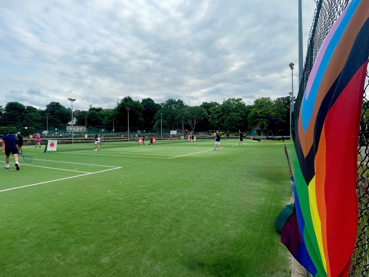 A view of multiple tennis courts with people playing on them. At the side of the image, on fencing at the back of the courts, a brighly coloured flag is attached to fencing