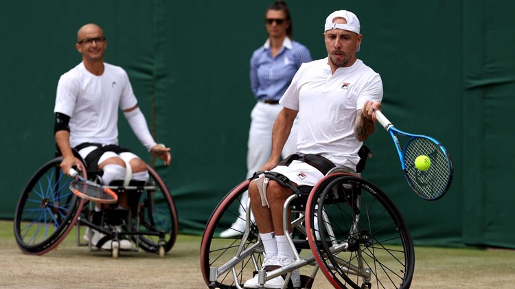 Andy Lapthorne preparing to hit a forehand on court while Guy Sasson is sat behind him