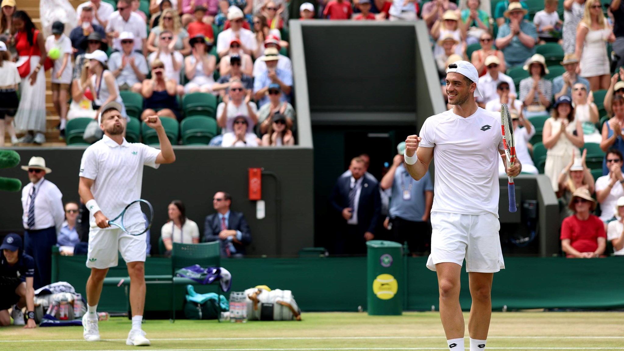 Julian Cash pumping his fist and racket in celebration while Lloyd Glasspool is behind him raising his fist in celebration in front of the Wimbledon crowd