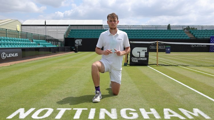 A man wearing white clothing kneels down on a grass tennis court, with the word 'Nottingham' written on the floor in front of him. A tennis net is visible over his left shoulder, and empty stands are situated behind and to the right of the man