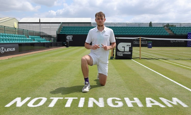 A man wearing white clothing kneels down on a grass tennis court, with the word 'Nottingham' written on the floor in front of him. A tennis net is visible over his left shoulder, and empty stands are situated behind and to the right of the man