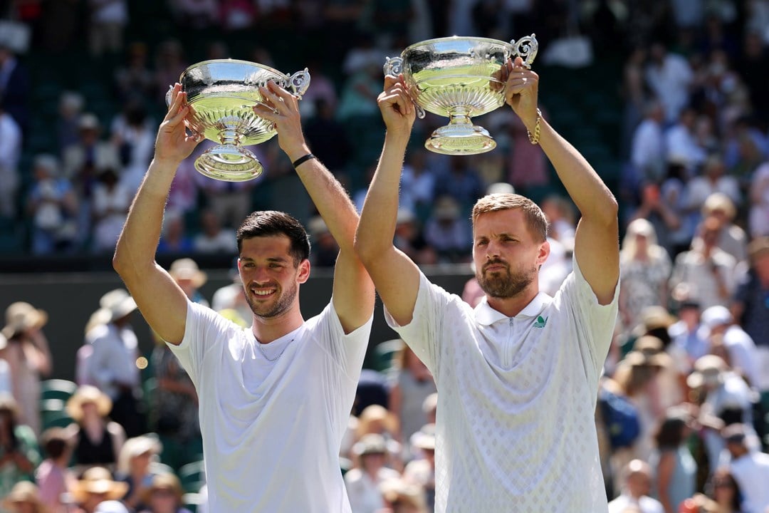 Julian Cash and Lloyd Glasspool smiling while holding up the Wimbledon men's doubles trophies above their head in front of the crowd