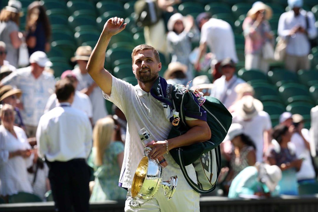 Lloyd Glasspool raising his hand to wave to the Wimbledon crowd, towel over his shoulder, holding onto his men's doubles trophy