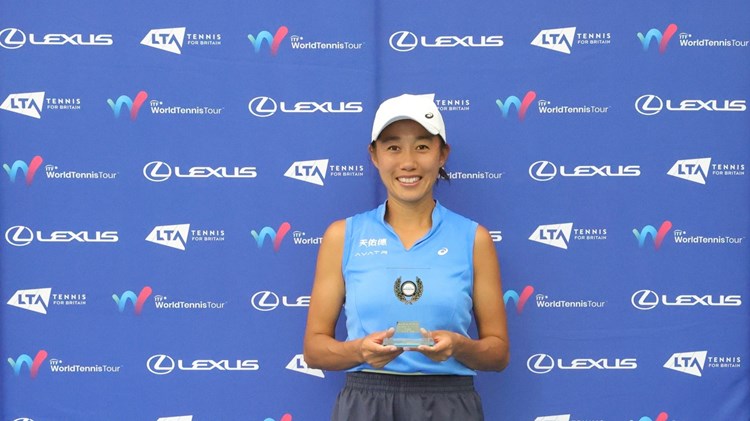 A woman wearing a blue sleeveless shirt, grey shorts and a white baseball cap stands in front of a blue background holidng a small glass trophy. Above her on the background read the words, 'Lexus British Pro Series'