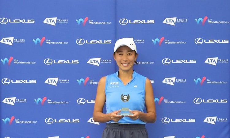A woman wearing a blue sleeveless shirt, grey shorts and a white baseball cap stands in front of a blue background holidng a small glass trophy. Above her on the background read the words, 'Lexus British Pro Series'