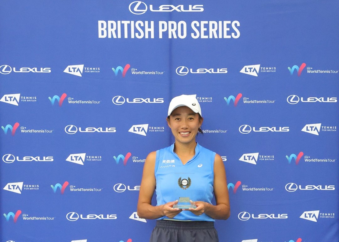 A woman wearing a blue sleeveless shirt, grey shorts and a white baseball cap stands in front of a blue background holidng a small glass trophy. Above her on the background read the words, 'Lexus British Pro Series'