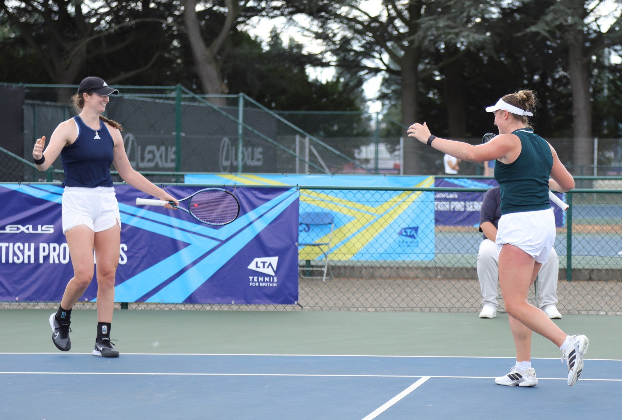 Two women on a tennis court smile facing each oither with their arms aloft.