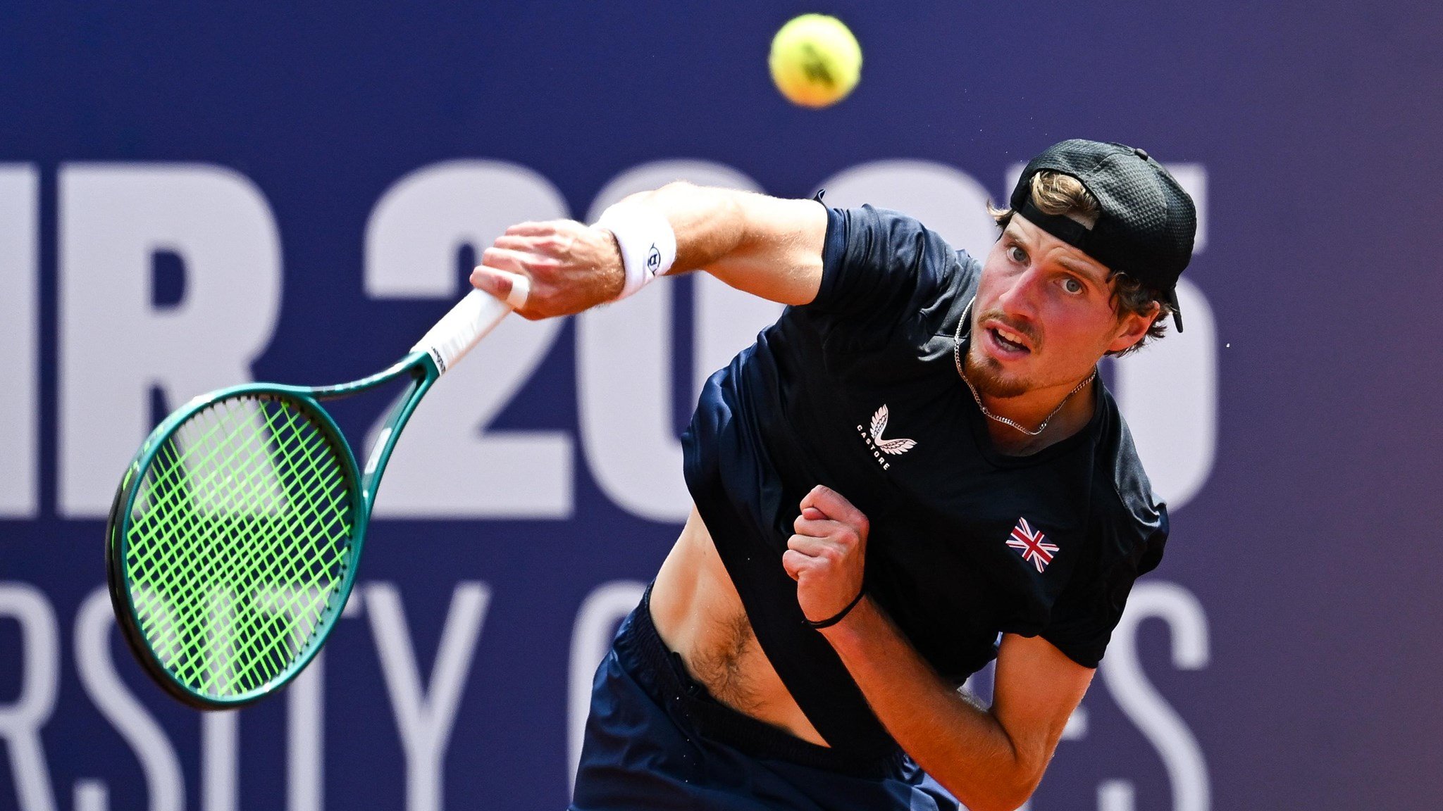 A man wearing a black cap and a dark blue shirt with the British flag on the left breast hits a tennis ball with a slightly strained expression on his face