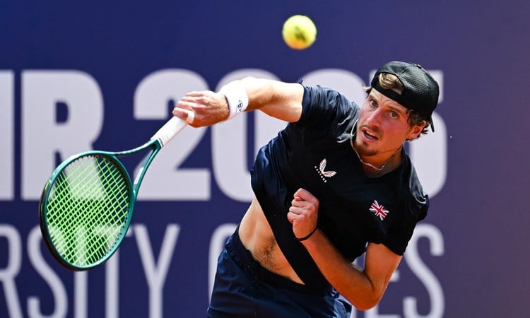 A man wearing a black cap and a dark blue shirt with the British flag on the left breast hits a tennis ball with a slightly strained expression on his face