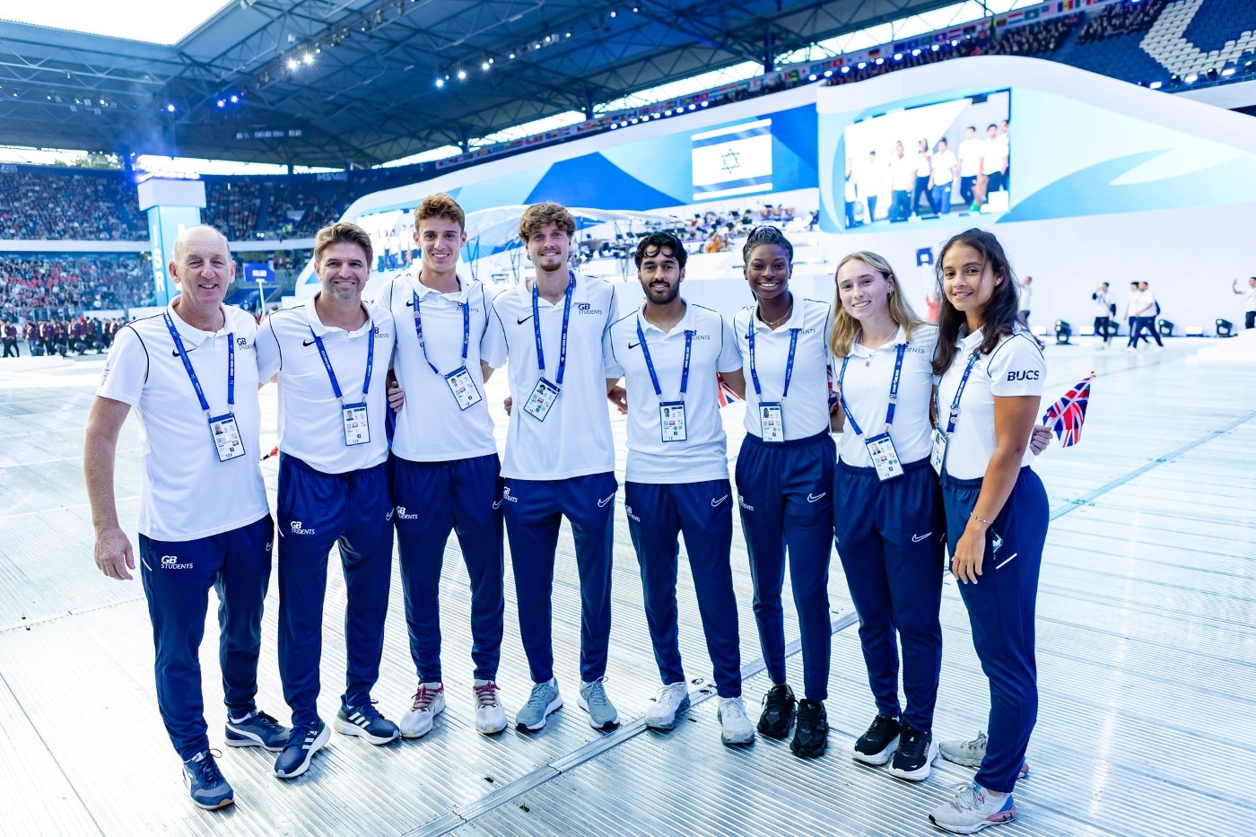 A group of eight people, five men and three women, all smile and are wearing white t-shirts and dark blue tracksuit bottoms