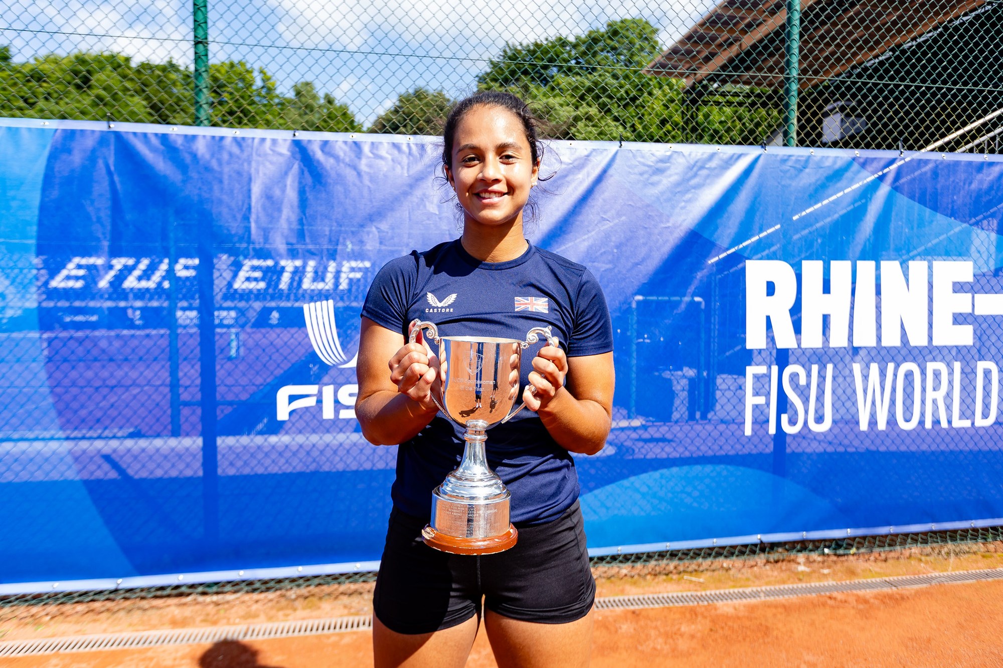 A woman stands similing, holding a large silver trophy. She is wearing a dark blue shirt with a British flag on the left breast, and black shorts, with a blue banner attached to a fence behind her