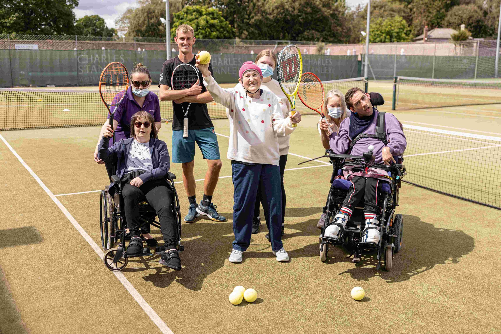 A group of sensory tennis participants holding up their rackets in front of the net and smiling.