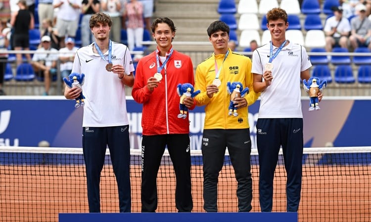 Four men wearing, from left to right, white, red, yellow and white sports tops, all showing of medals hung around their necks. A tennis net is directly behind them, and crowds are visible in stands in the background