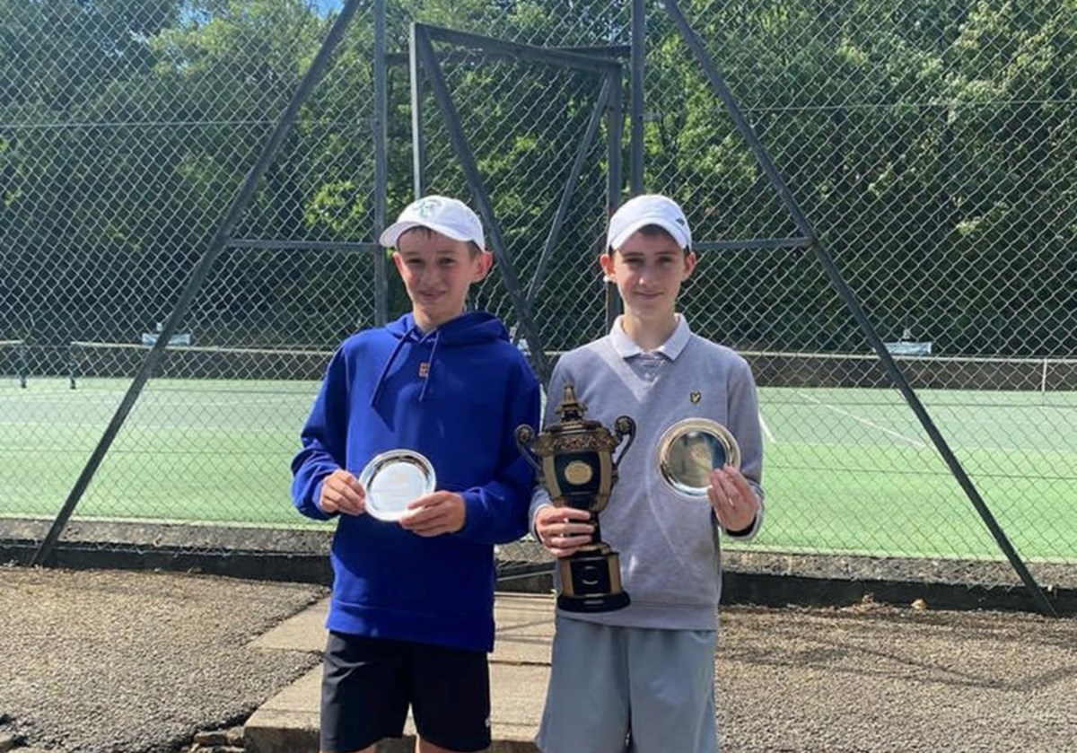Two boys pose with their trophies beside a tennis court