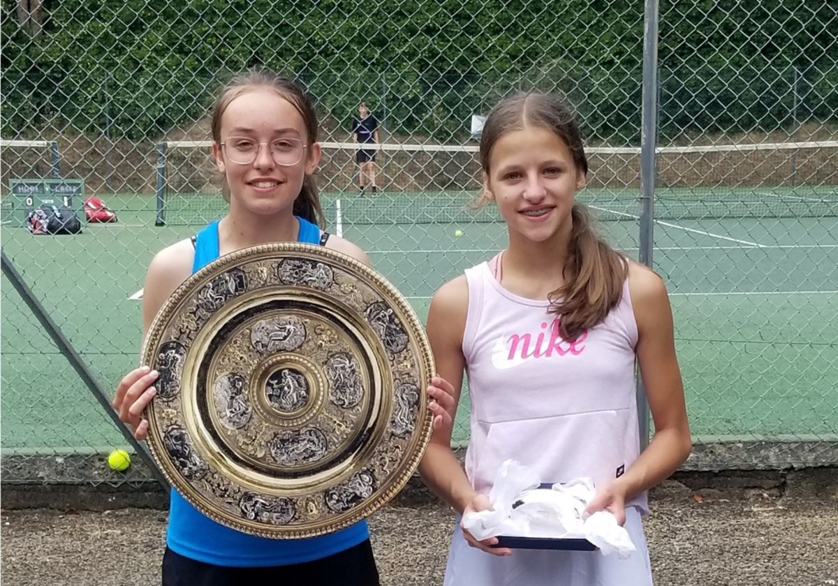 Two girls pose with their tennis trophies beside tennis court